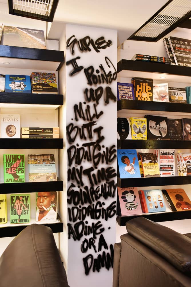 Black shelving stacked with hip-hop and Nigerian titles beside a hand-sprayed lyric column